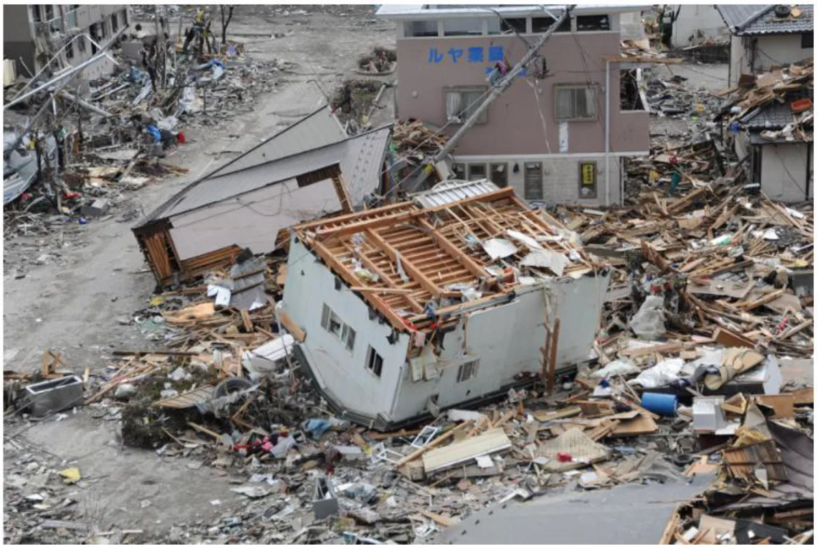 東日本大震災の津波で破壊された市街地の空撮写真。基礎から流されて傾いた木造家屋が道路の上に横たわり、周囲一面に建材や家財が散乱している。